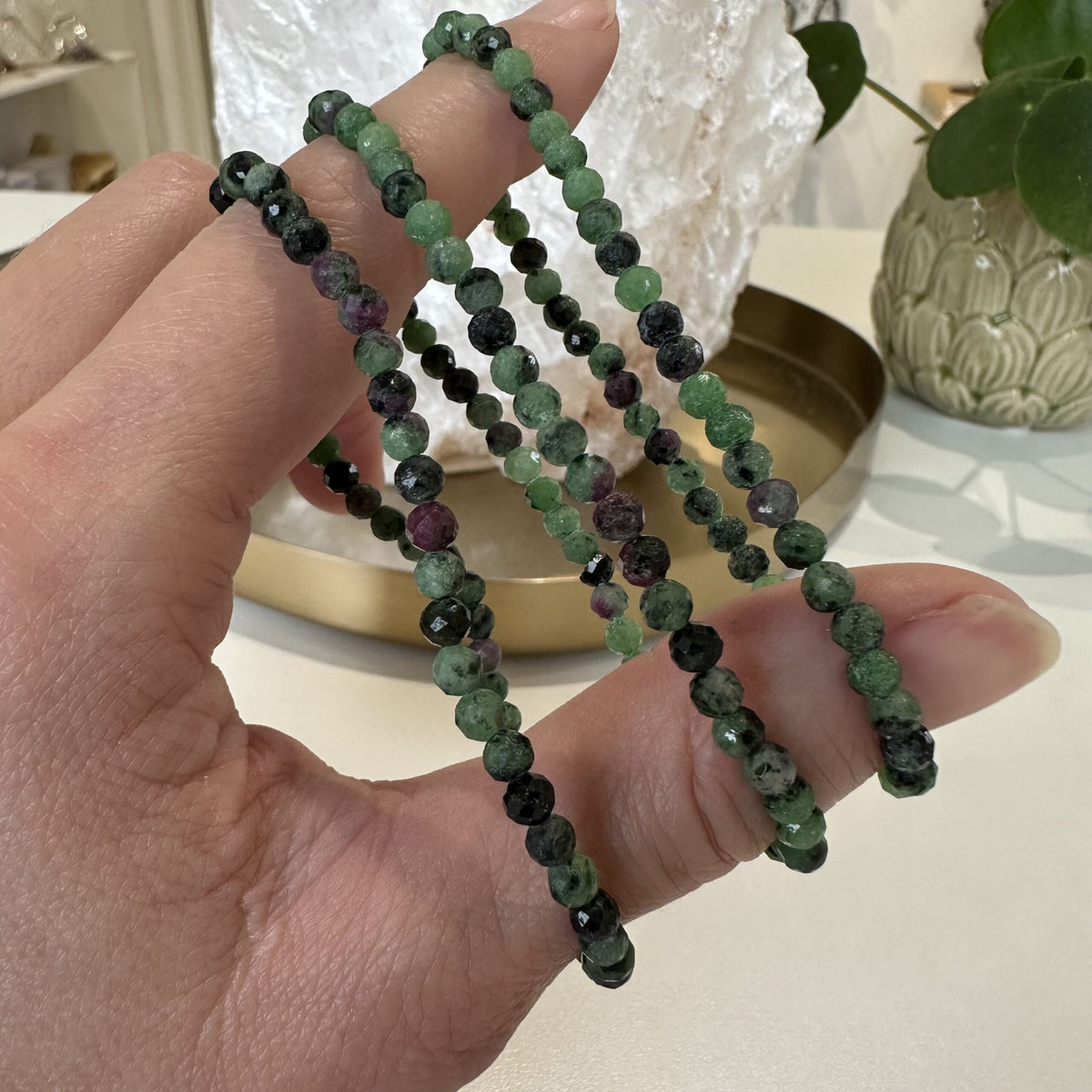 Hånd holder Zoisit Rubin armbånd med grønne og lilla sten, i et rum med planter og dekorative genstande.