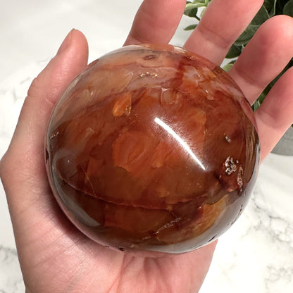 En hånd holder en glat, poleret karneol kugle med dybe orange og røde nuancer.