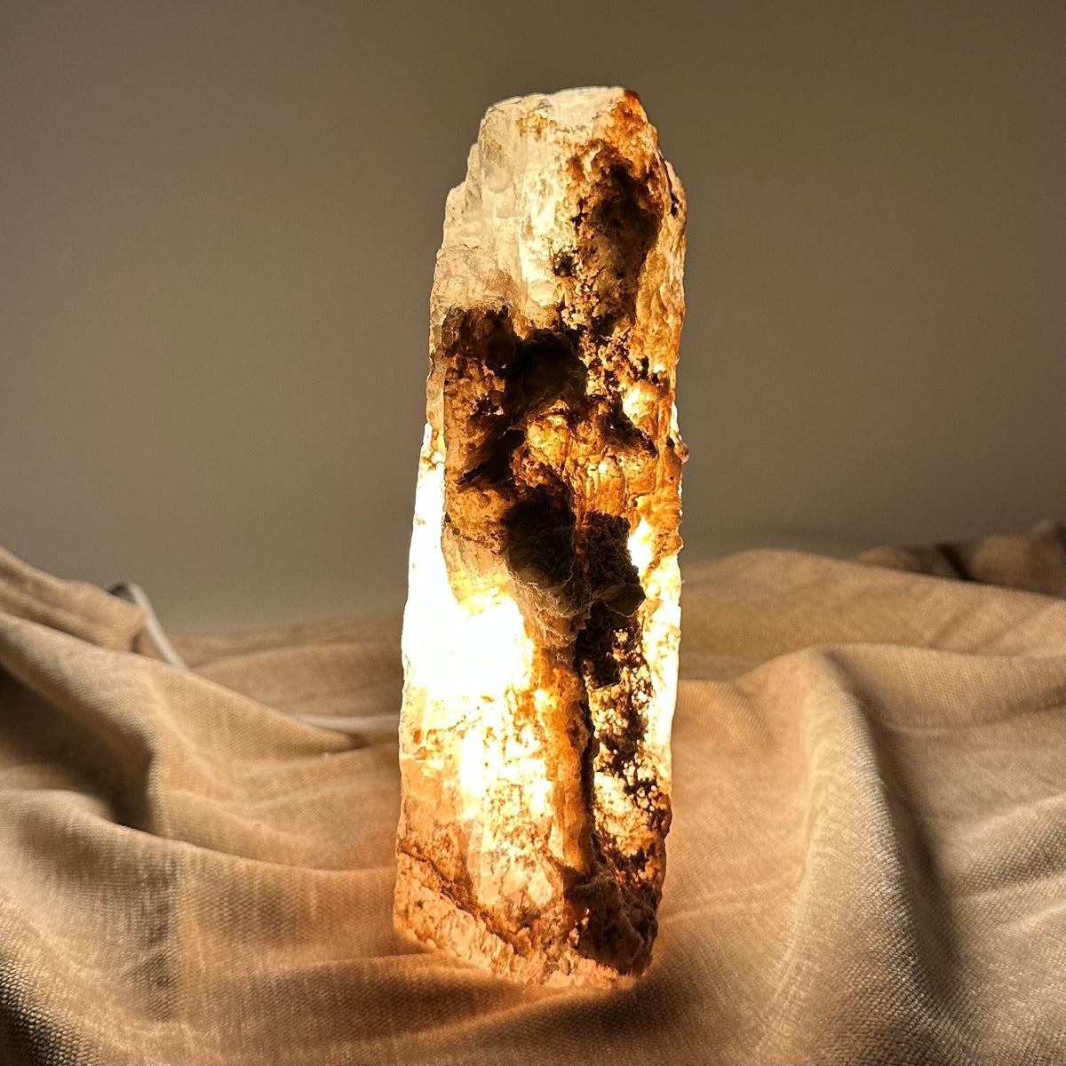 Mexicansk Selenit lampe nr 51 lyser op på en beige overflade, med sine naturlige, rå detaljer tydeligt synlige.