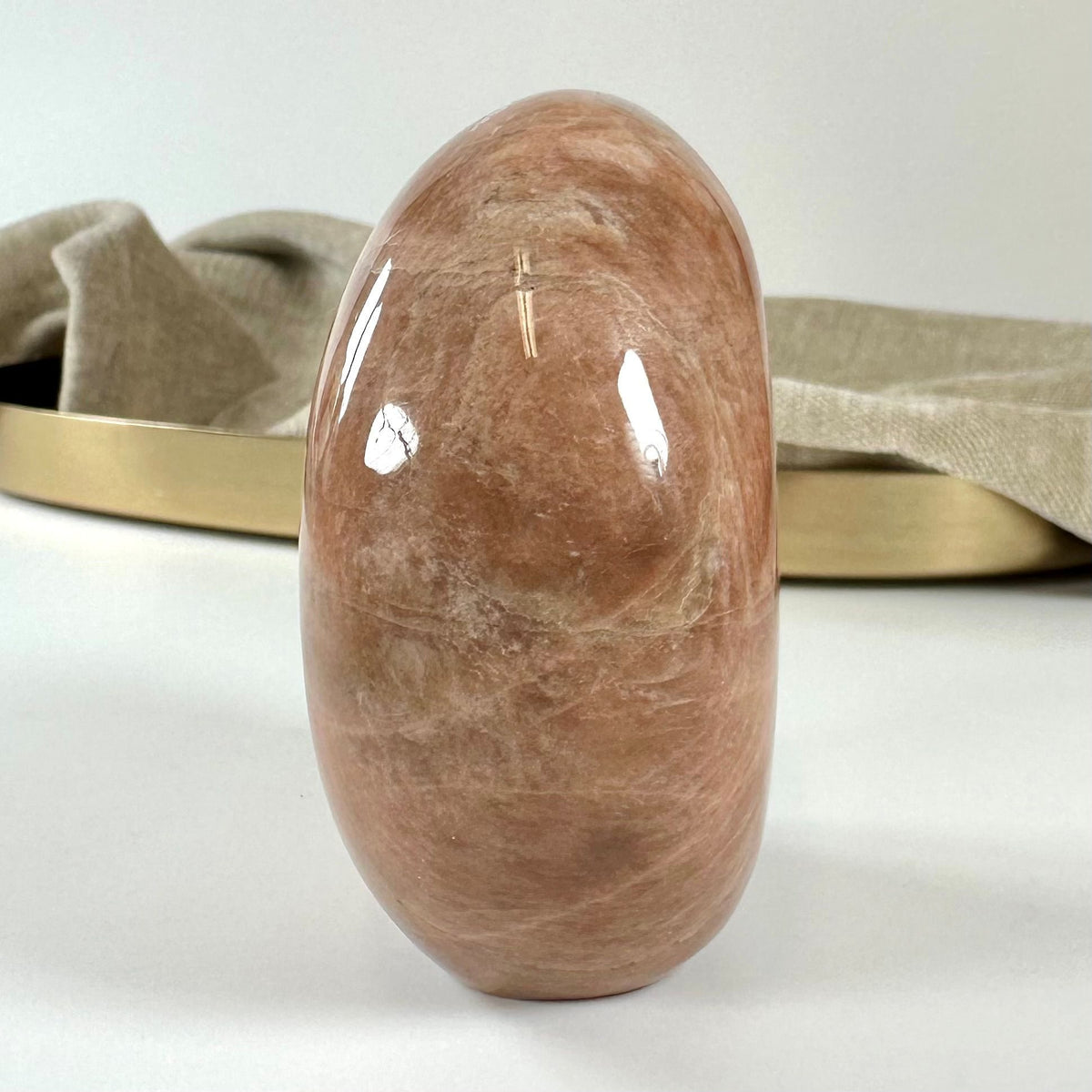 En glat fersken månesten på en lys overflade, baggrund med beige stof og en guldbakke.