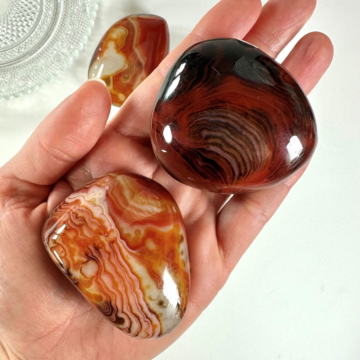Hånd med to Sardonyx palmstones i røde og orange nuancer, placeret på hvid baggrund.