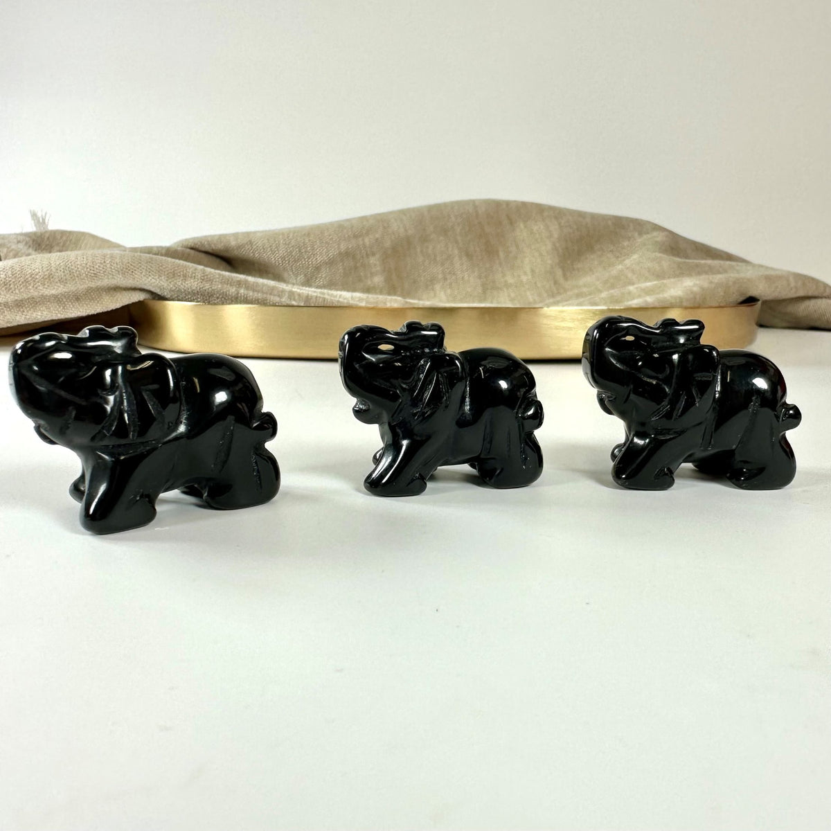 Tre små sorte obsidian elefanter på en hvid overflade med en beige klud og et metalfad i baggrunden.