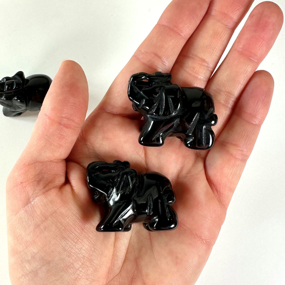 Hånd holder to små, skinnende sorte obsidian elefantfigurer; en tredje ses i baggrunden på en lys baggrund.