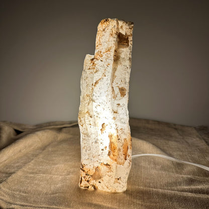 Mexicansk Selenit lampe nr. 54 er en håndskåret stemningslampe af naturlig selenitkrystal med unikke strukturer og varme jordfarver. Lampen udsender et blødt, dæmpbart lys, perfekt til meditation, ro og afslapning. Den står på et neutralt underlag og kaster et varmt skær på omgivelserne. Et unikt stykke belysning, der kombinerer naturens skønhed med spirituel energi.