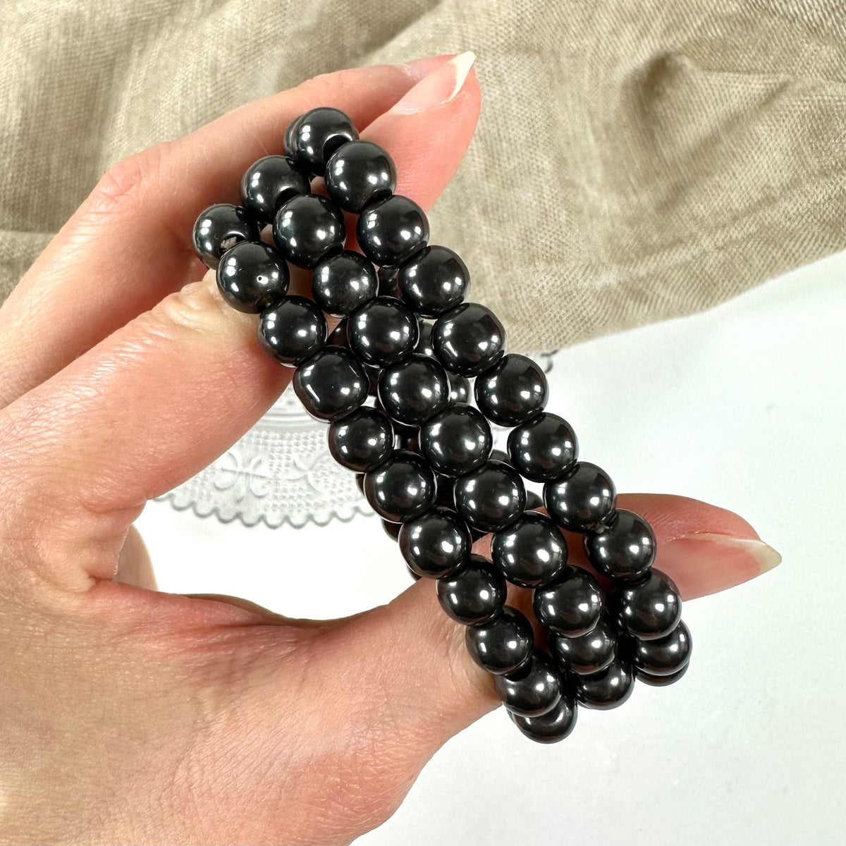Hånden holder flere skinnende, sorte hæmatit-armbånd mod en beigefarvet baggrund.