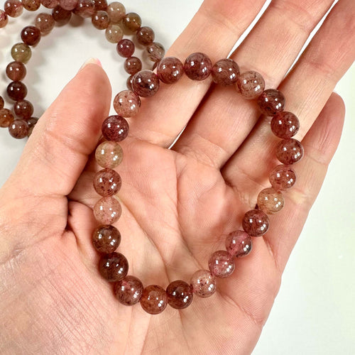 En hånd holder et jordbærkvarts armbånd med glatte, rødbrune perler.