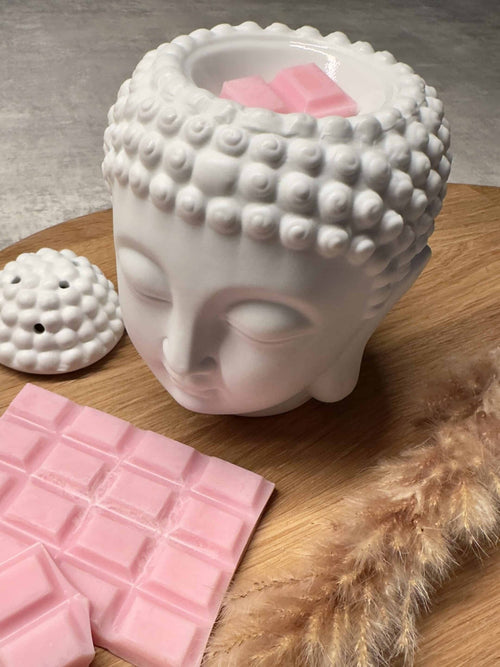 Voksbrænder med Buddha i hvid keramik og lyserøde voksstykker på en træplade, sammen med tørrede blomster.