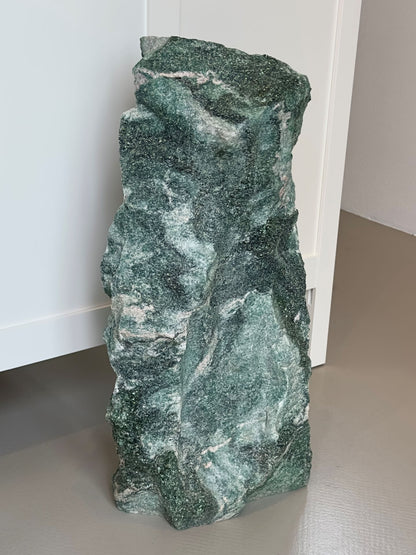 Fuchsit unika sten i varierende grønne toner står på gulvet ved en hvid væg.