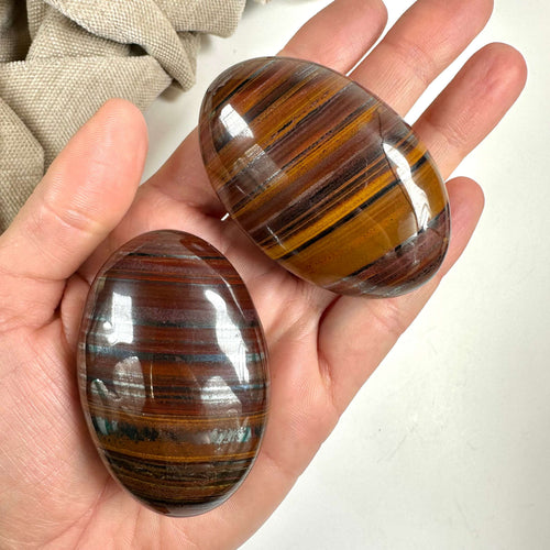 Hånd holder to polerede okse- og tigerøje palmstones med brune og gyldne striber.
