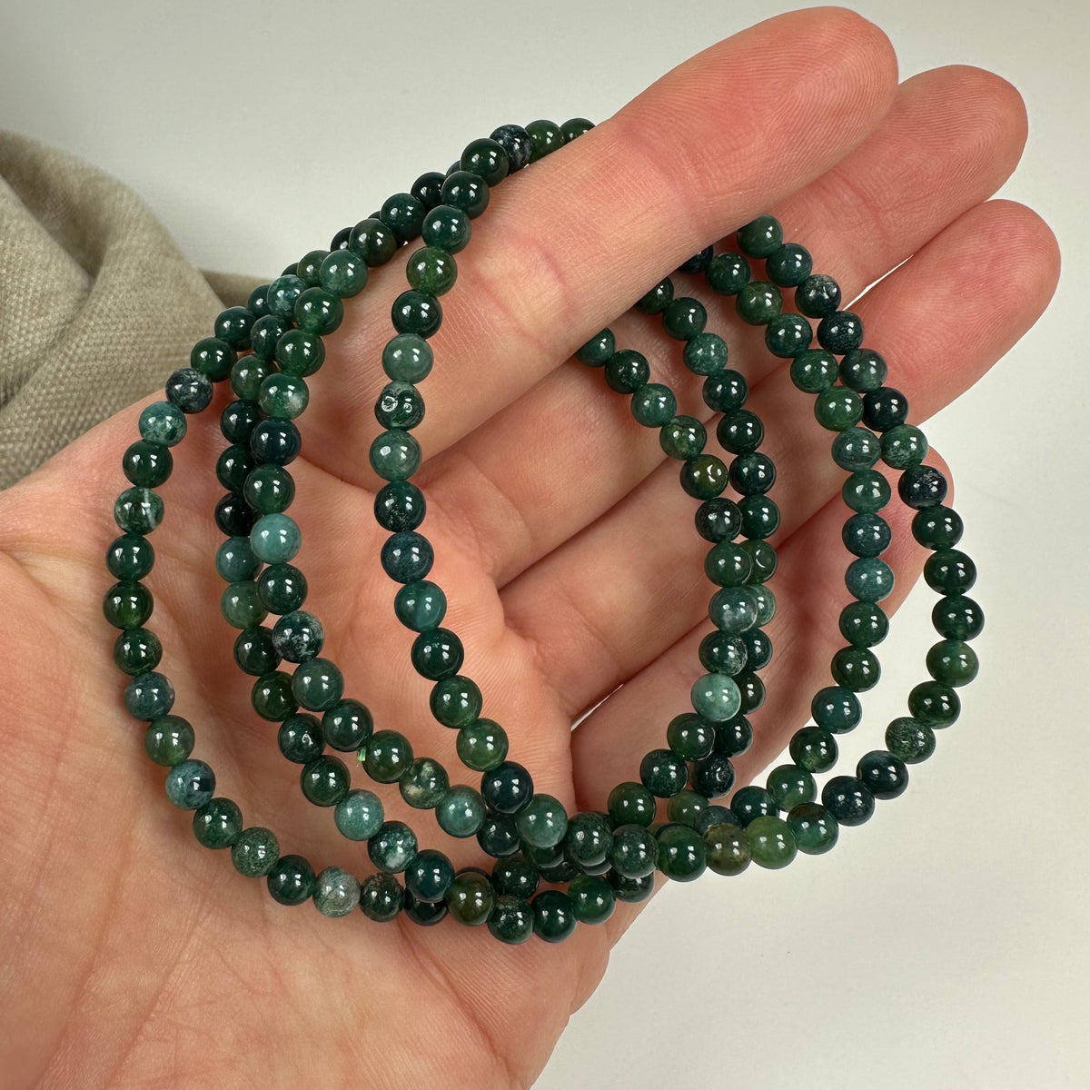 Hånd holder flere grønne Mos Agat armbånd på en beige baggrund.