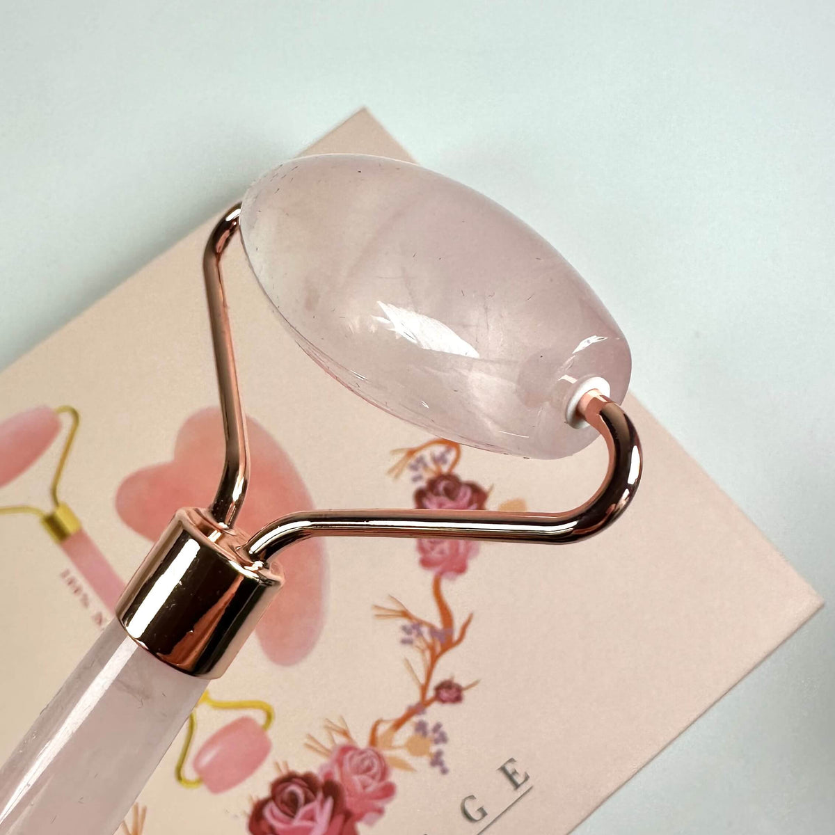 Gua sha sæt med rosakvarts massage-rulle på pastelfarvet baggrund med blomsterdetaljer.