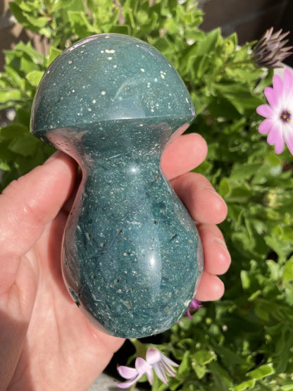 En hånd holder en Jaspis hav paddehat nr4 foran grønne planter og en lyserød blomst.