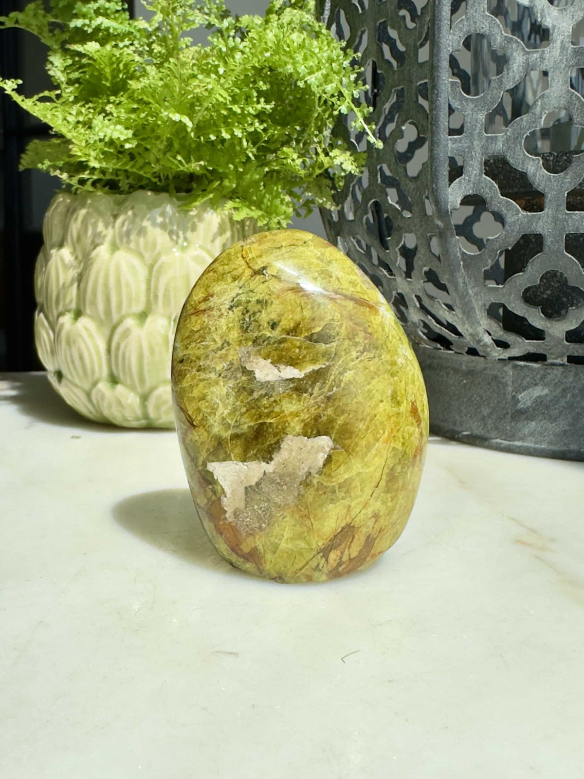 Grøn opal i fri form på bord, omgivet af en dekorativ plante og lantern.