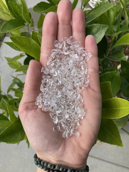 Håndfuld Bjergkrystal mini på 100 gram, holdt i hånden med grønne blade i baggrunden.